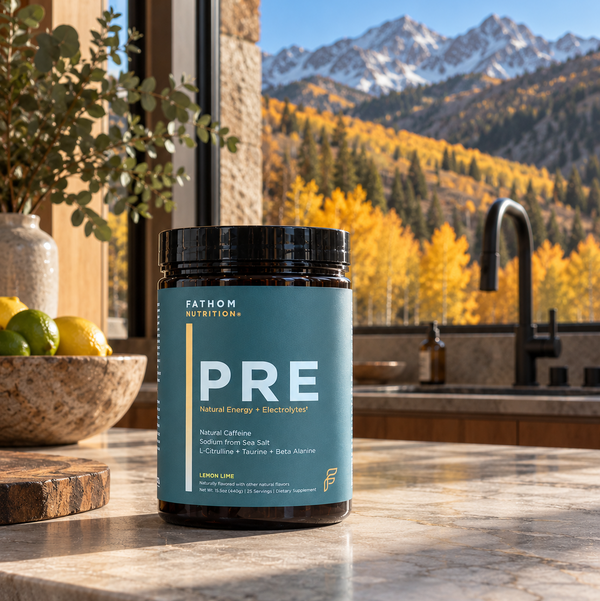 A tub of Fathom Nutrition PRE supplement sits on a marble kitchen counter with a mountain and autumn forest view in the background.