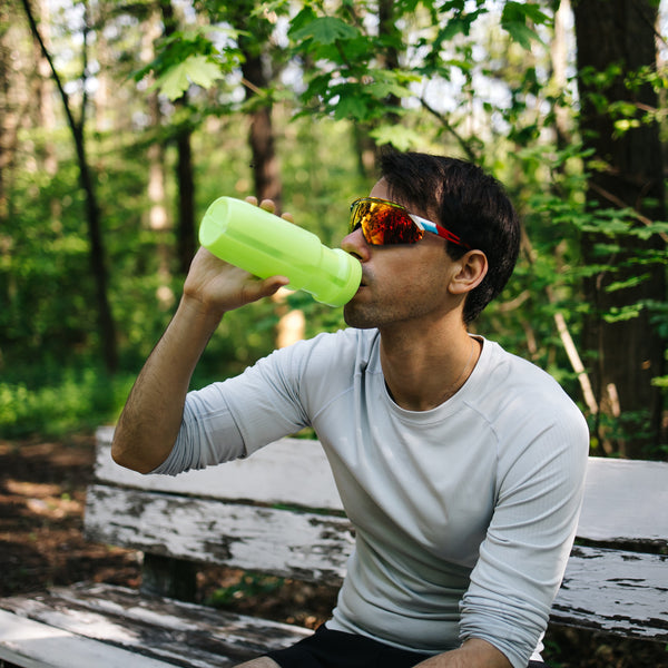 A man wearing a white long-sleeve shirt and orange-tinted sunglasses sits on a weathered wooden bench in a forest, drinking from a bright neon green water bottle.