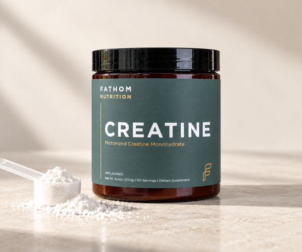 A jar of Fathom Nutrition Creatine Micronized Creatine Monohydrate with a dark green label and black lid, shown on a light stone surface with a scoop of white powder beside it.