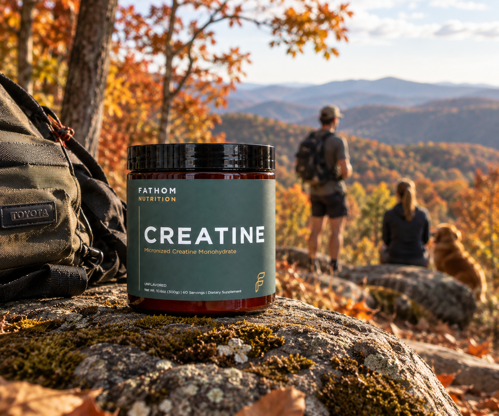 A jar of Fathom Nutrition Creatine Micronized Creatine Monohydrate with a dark green label and black lid, shown on a light stone surface with a scoop of white powder beside it.